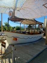A small outdoor restaurant stand designed to look like a boat on a wooden deck, featuring a sign with the text 'Fish in the Fingers'. The stand is covered by a camo net providing shade, and surrounded by greenery and mountain views.