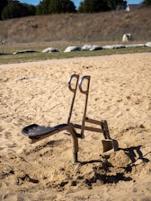 An outdoor exercise machine is set in a sandy area. It appears to be designed for digging or leg exercises, with a seat and handles. The background features a natural landscape with dry grass and rocks under a clear sky.