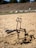 An outdoor exercise machine is set in a sandy area. It appears to be designed for digging or leg exercises, with a seat and handles. The background features a natural landscape with dry grass and rocks under a clear sky.