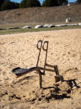 An outdoor exercise machine is set in a sandy area. It appears to be designed for digging or leg exercises, with a seat and handles. The background features a natural landscape with dry grass and rocks under a clear sky.