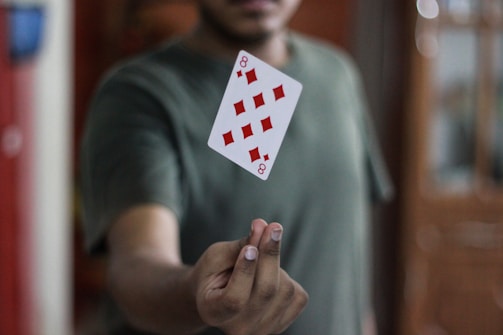 A person holds a playing card between their fingers, showcasing the eight of diamonds. The background is blurred, emphasizing the focus on the card and hand.