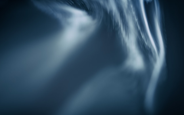 Abstract close-up featuring smooth, flowing lines with a blurred effect, creating a sense of motion. The color palette is predominantly dark with shades of blue and hints of white.
