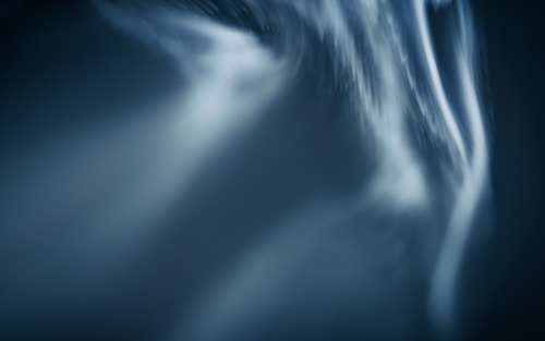 Abstract close-up featuring smooth, flowing lines with a blurred effect, creating a sense of motion. The color palette is predominantly dark with shades of blue and hints of white.