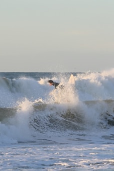 A surfer skillfully navigates a large, powerful wave in the ocean, with white foam and spray enveloping the scene. The sky is clear and a subtle gradient of light blue and soft yellow.