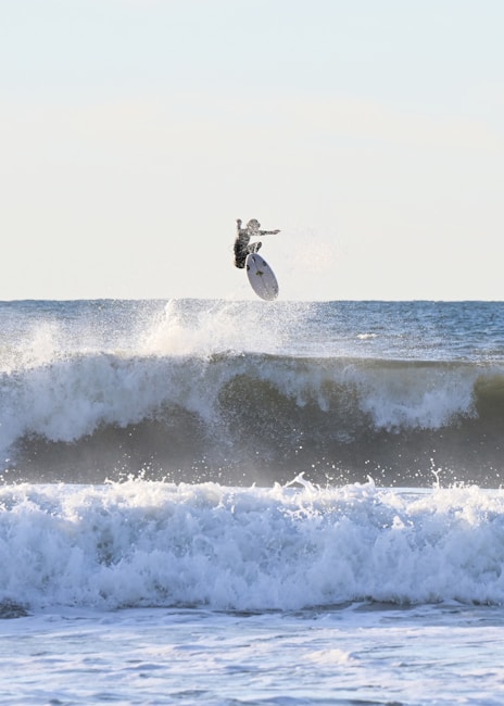 A surfer is captured mid-air above a large wave, performing a trick with their surfboard nearly vertical. The ocean appears turbulent with multiple waves visible, and the sky is clear with no clouds. The action takes place during daylight, evidenced by the brightness of the scene.
