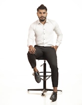 A man with a beard and styled hair is sitting on a stool. He is wearing a white button-up shirt, black pants, and black shoes. His posture is relaxed with one hand resting on his leg and the other in his pocket. The background is plain and white, giving a minimalistic feel.