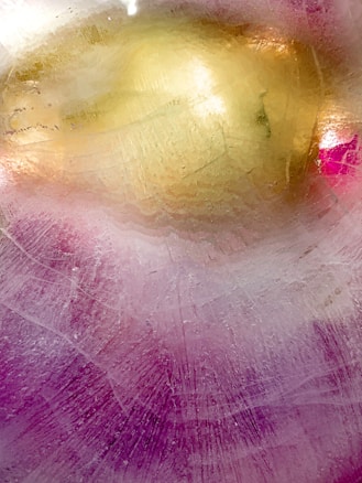 A textured surface with a rich blend of colors, predominantly featuring a golden area blended with purple and hints of pink. The surface appears to have a frosty or icy appearance, with visible striations and scratches adding depth.