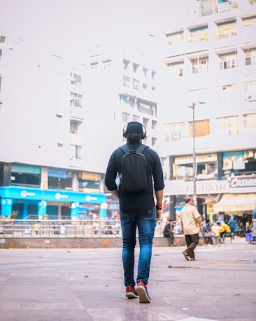 A person wearing headphones and a backpack walks on a city street. The background features tall buildings and shops, with a few people visible in the distance. The scene has an urban, busy atmosphere.