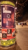 A cylindrical outdoor advertisement pillar displays multiple posters for upcoming music events. The prominent poster advertises Sr. Chinarro performing at Sala Wolf on February 10. Other posters announce concerts by Kevin Johansen on February 1, Christina Rosenvinge on February 4, and Núria Graham on February 17. The scene is set on a dimly lit street at night, with blurry figures and streetlights in the background.