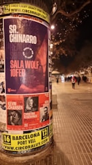 A cylindrical outdoor advertisement pillar displays multiple posters for upcoming music events. The prominent poster advertises Sr. Chinarro performing at Sala Wolf on February 10. Other posters announce concerts by Kevin Johansen on February 1, Christina Rosenvinge on February 4, and Núria Graham on February 17. The scene is set on a dimly lit street at night, with blurry figures and streetlights in the background.