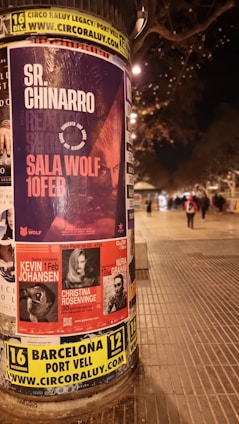 A cylindrical outdoor advertisement pillar displays multiple posters for upcoming music events. The prominent poster advertises Sr. Chinarro performing at Sala Wolf on February 10. Other posters announce concerts by Kevin Johansen on February 1, Christina Rosenvinge on February 4, and Núria Graham on February 17. The scene is set on a dimly lit street at night, with blurry figures and streetlights in the background.