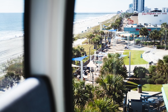 A coastal scene featuring a beach with gentle waves and sandy shores running parallel to a busy boardwalk. Palm trees line the walkway, and there are several tourists enjoying the area. Buildings and storefronts in the background include a sign indicating 'Gift Cove.' The area is bathed in bright sunlight, creating a vibrant and lively atmosphere.
