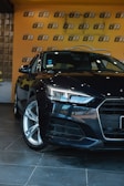 A sleek, black car is positioned in an indoor showroom with a glossy tiled floor. The background features a bright orange wall with a repeating black and white logo pattern. The car's headlights are prominently featured, emphasizing its modern design.