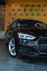 A sleek, black car is positioned in an indoor showroom with a glossy tiled floor. The background features a bright orange wall with a repeating black and white logo pattern. The car's headlights are prominently featured, emphasizing its modern design.