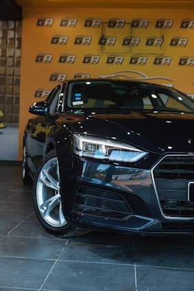 A sleek, black car is positioned in an indoor showroom with a glossy tiled floor. The background features a bright orange wall with a repeating black and white logo pattern. The car's headlights are prominently featured, emphasizing its modern design.