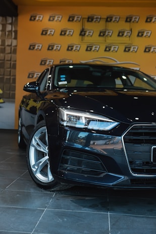 A sleek, black car is positioned in an indoor showroom with a glossy tiled floor. The background features a bright orange wall with a repeating black and white logo pattern. The car's headlights are prominently featured, emphasizing its modern design.