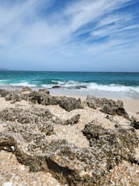 Rock formations on a sandy beach with waves crashing in the turquoise sea under a partly cloudy sky.