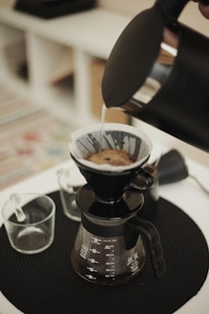 A pour-over coffee setup with hot water being poured from a kettle over coffee grounds contained in a filter. The black coffee dripper and glass carafe sit on a black mat. Two glass cups are placed nearby.