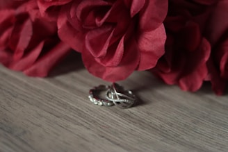 A pair of intertwined silver rings with small embedded gems is placed on a wooden surface. Above the rings, a bouquet of red roses provides a striking contrast with its rich color.