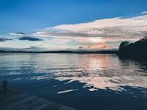 The photograph captures a serene lake scene during either sunrise or sunset, with calm waters reflecting the colors of the sky. Wispy clouds stretch across the sky, merging with soft hues of orange and pink as the sun is partially hidden. Trees line the horizon, creating a dark silhouette against the vibrant sky. A wooden dock extends into the foreground, adding a rustic touch to the peaceful setting.