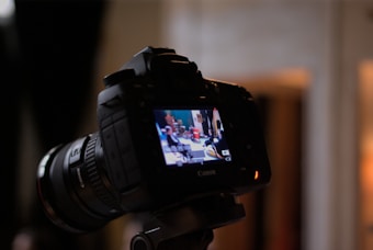 A digital camera is positioned on a tripod, capturing a scene displayed on its LCD screen. The screen shows a brightly lit indoor setting with several indistinct figures or objects in the background. The camera is in focus, while the background is blurred.