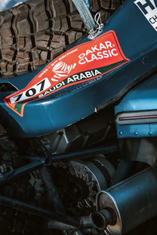 A close-up view of mechanical components of a vehicle featuring a prominent tire and engine parts. The vehicle sports a sticker with 'Dakar Classic' and 'Saudi Arabia' along with the number 707, suggesting participation in a rally event.