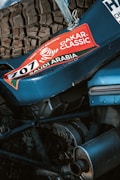 A close-up view of mechanical components of a vehicle featuring a prominent tire and engine parts. The vehicle sports a sticker with 'Dakar Classic' and 'Saudi Arabia' along with the number 707, suggesting participation in a rally event.