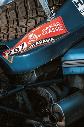 A close-up view of mechanical components of a vehicle featuring a prominent tire and engine parts. The vehicle sports a sticker with 'Dakar Classic' and 'Saudi Arabia' along with the number 707, suggesting participation in a rally event.