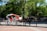 A horse-drawn carriage with a white and red design is parked along a path in a park. The carriage has large white wheels and a red interior. The background is filled with lush, green trees, and several people can be seen casually walking and standing nearby, enjoying a sunny day.