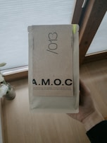 A hand is holding a beige package labeled with the letters A.M.O.C. in bold. The package features additional text indicating it contains specialty coffee. The background includes a wooden floor and partially visible window with blinds.