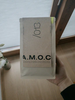 Hands holding a box of Brewmosaic coffee blends with a cozy kitchen setting in the background.