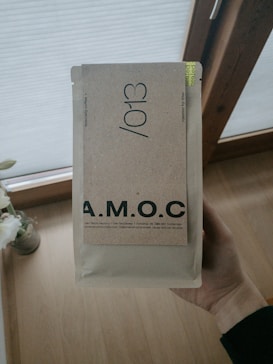 A hand is holding a beige package labeled with the letters A.M.O.C. in bold. The package features additional text indicating it contains specialty coffee. The background includes a wooden floor and partially visible window with blinds.
