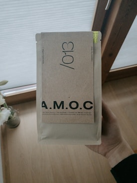 A hand is holding a beige package labeled with the letters A.M.O.C. in bold. The package features additional text indicating it contains specialty coffee. The background includes a wooden floor and partially visible window with blinds.