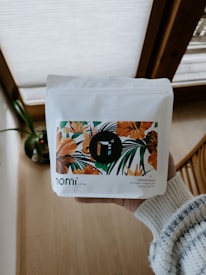 A person is holding a white package of coffee with floral artwork featuring orange and green leaves. The words 'nomi coffee' and 'KERAMO VILLAGE' are visible. The background includes a wooden floor and some natural light from a window.
