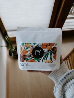 A person is holding a white package of coffee with floral artwork featuring orange and green leaves. The words 'nomi coffee' and 'KERAMO VILLAGE' are visible. The background includes a wooden floor and some natural light from a window.