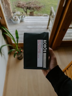 A person's hand is holding a black package with a label that reads 'LOT61'. The background features a wooden deck with some plant pots and a green lawn visible outside a glass door. The setting seems to be inside a cozy room with wooden floors and furniture.