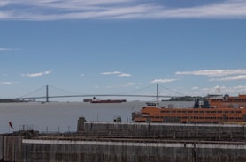 A ferry labeled 'Staten Island Ferry' is docked near the wooden pier in the foreground. In the background, a large suspension bridge stretches across the waterway, with a cargo ship passing underneath. The sky is partly cloudy with patches of blue visible.