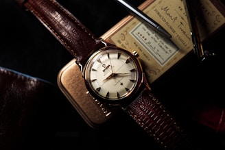 A vintage wristwatch with a brown leather strap is prominently displayed on top of a tobacco tin. The watch face is cream-colored with intricate, gold-toned hour markers and hands. The background contains a textured object and a pen, adding to the classic and sophisticated aesthetic.