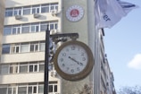 A large vintage-style clock is mounted on a pole in front of a building with multiple windows. Air conditioning units are visible on the building's exterior. A round red emblem with legal symbols is placed on the wall. Two flags are flying next to the clock, partially readable due to their movement.