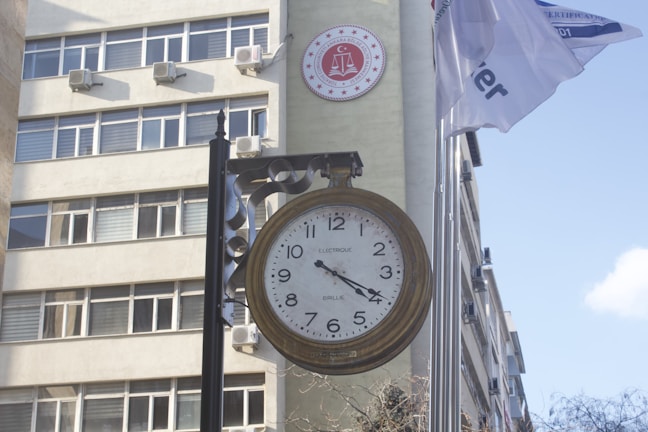A large vintage-style clock is mounted on a pole in front of a building with multiple windows. Air conditioning units are visible on the building's exterior. A round red emblem with legal symbols is placed on the wall. Two flags are flying next to the clock, partially readable due to their movement.
