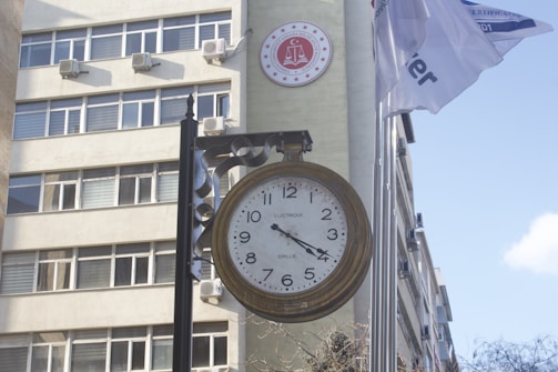 A large vintage-style clock is mounted on a pole in front of a building with multiple windows. Air conditioning units are visible on the building's exterior. A round red emblem with legal symbols is placed on the wall. Two flags are flying next to the clock, partially readable due to their movement.