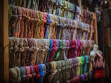 A display of numerous colorful scarves is organized neatly on wooden shelves. The scarves are arranged in rows, showcasing a variety of patterns and fabrics. A mannequin dressed in a sleeveless dress is positioned to the right side of the image.