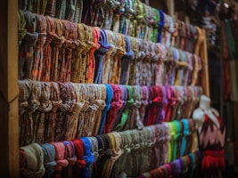 A display of numerous colorful scarves is organized neatly on wooden shelves. The scarves are arranged in rows, showcasing a variety of patterns and fabrics. A mannequin dressed in a sleeveless dress is positioned to the right side of the image.