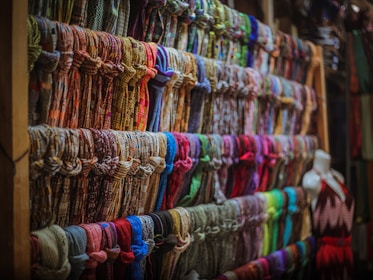 A display of numerous colorful scarves is organized neatly on wooden shelves. The scarves are arranged in rows, showcasing a variety of patterns and fabrics. A mannequin dressed in a sleeveless dress is positioned to the right side of the image.