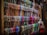A display of numerous colorful scarves is organized neatly on wooden shelves. The scarves are arranged in rows, showcasing a variety of patterns and fabrics. A mannequin dressed in a sleeveless dress is positioned to the right side of the image.