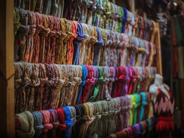 A display of numerous colorful scarves is organized neatly on wooden shelves. The scarves are arranged in rows, showcasing a variety of patterns and fabrics. A mannequin dressed in a sleeveless dress is positioned to the right side of the image.