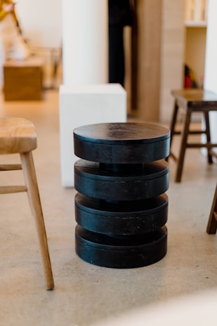 A modern interior space with a black, cylindrical, multi-layered stool or side table in the center. Surrounding it are wooden chairs and light-colored furniture pieces, creating a minimalist and contemporary vibe. The flooring is of a neutral tone, complementing the light walls and furnishings.