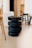 A modern interior scene featuring a stack of black, cylindrical stools placed centrally. The stools are surrounded by an assortment of other wooden chairs and minimalistic furniture in a softly lit room with a smooth floor and neutral-colored walls.