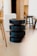 A modern interior scene featuring a stack of black, cylindrical stools placed centrally. The stools are surrounded by an assortment of other wooden chairs and minimalistic furniture in a softly lit room with a smooth floor and neutral-colored walls.