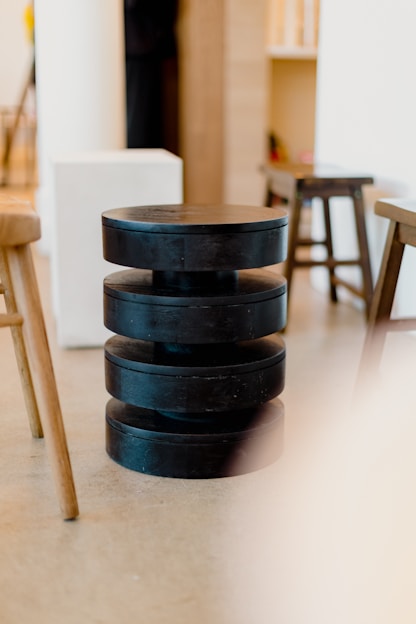 A modern interior scene featuring a stack of black, cylindrical stools placed centrally. The stools are surrounded by an assortment of other wooden chairs and minimalistic furniture in a softly lit room with a smooth floor and neutral-colored walls.